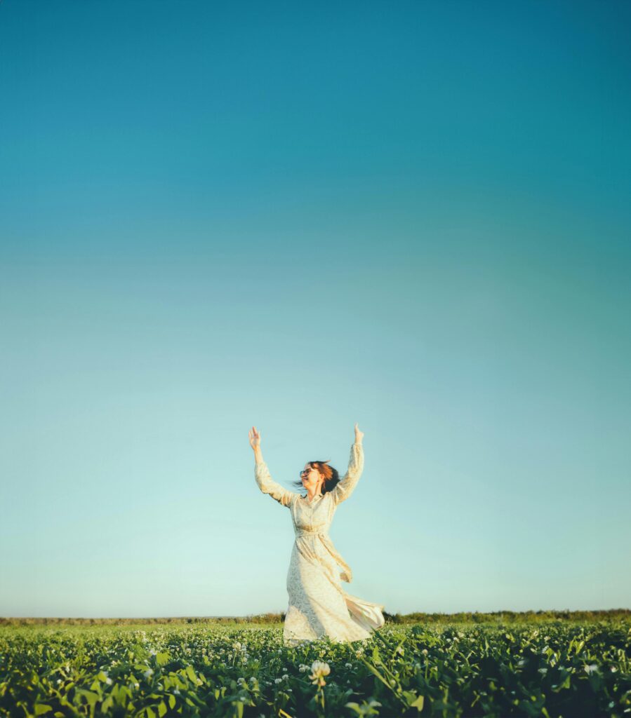 A joyful woman in a field with arms raised against a clear blue sky, depicting freedom and happiness.
