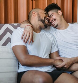 A joyful gay couple cuddling and smiling on a couch, showcasing affection and happiness.