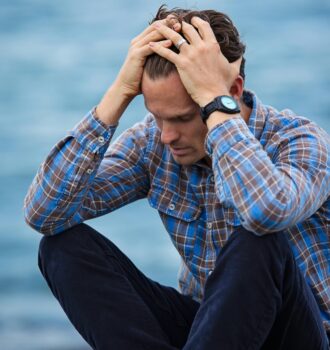 A man in a plaid shirt sits by the water looking distressed, symbolizing stress.