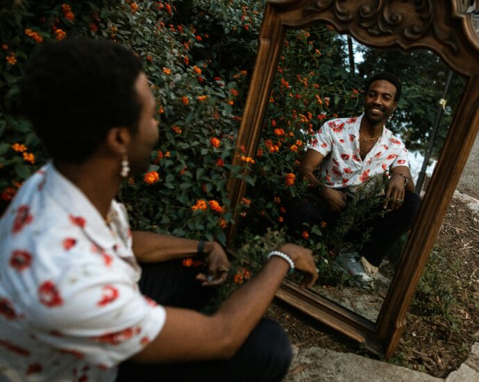 A man smiling at his reflection in a mirror surrounded by vibrant flowers outdoors.