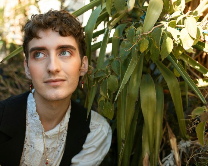 Young adult with delicate earrings poses among lush green plants in soft lighting.